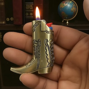 May include: A brass-colored cowboy boot lighter with a flame. The lighter is shaped like a boot with decorative details. The flame is orange and yellow. The lighter is held in a hand.