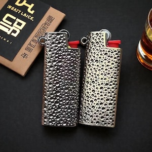 Op de afbeelding: Twee metalen aanstekers met een textuur, bubbelachtig ontwerp. Eén is zilver, de andere goud. Beide hebben een rode ontstekingsknop. Een kleine bruine doos met gouden tekst is zichtbaar op de achtergrond.