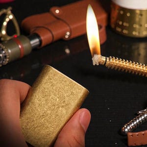 May include: A gold-colored rectangular lighter is held in a hand. A long, flexible, gold-colored lighter with a lit flame is in the background. Other items in the background include a brown leather case and a decorative gold-colored item.
