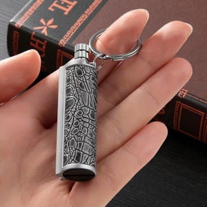 May include: A silver keychain lighter with a textured, crocodile-skin pattern. The lighter is attached to a silver key ring. The lighter is held in a person's hand, with a book in the background.