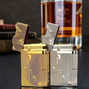 Puede incluir: Dos encendedores met&aacute;licos, uno dorado y otro plateado, con intrincados dise&ntilde;os grabados. Los encendedores est&aacute;n abiertos, revelando sus mecanismos internos. El encendedor dorado tiene un patr&oacute;n vertical, mientras que el plateado tiene un dise&ntilde;o ondulado. Un vaso de l&iacute;quido &aacute;mbar est&aacute; en el fondo.