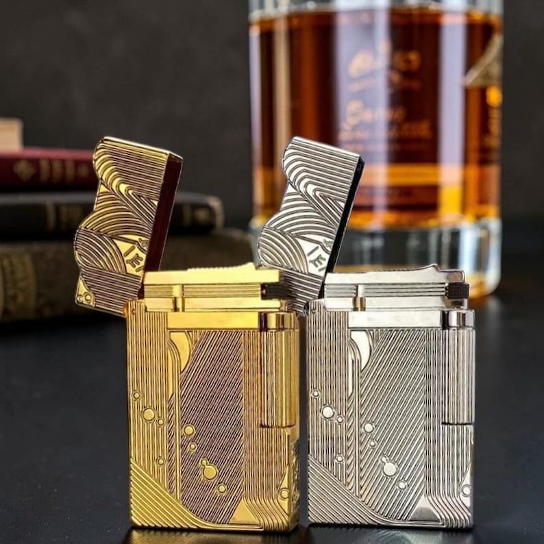 Puede incluir: Dos encendedores met&aacute;licos, uno dorado y otro plateado, con intrincados dise&ntilde;os grabados. Los encendedores est&aacute;n abiertos, revelando sus mecanismos internos. El encendedor dorado tiene un patr&oacute;n vertical, mientras que el plateado tiene un dise&ntilde;o ondulado. Un vaso de l&iacute;quido &aacute;mbar est&aacute; en el fondo.