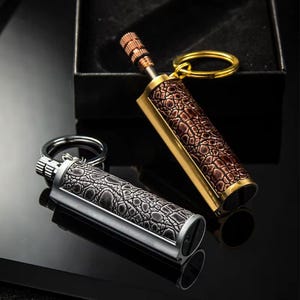 May include: Two metal keychain lighters. One is silver with a textured pattern, the other is gold with a brown, textured pattern. Both have a cylindrical body and a ring for keys. The lighters are on a reflective surface.