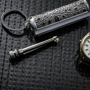 May include: A silver and black keychain lighter with a crocodile skin pattern. A separate metal striker and a gold wristwatch with a black strap are also visible. The items are on a textured black surface.