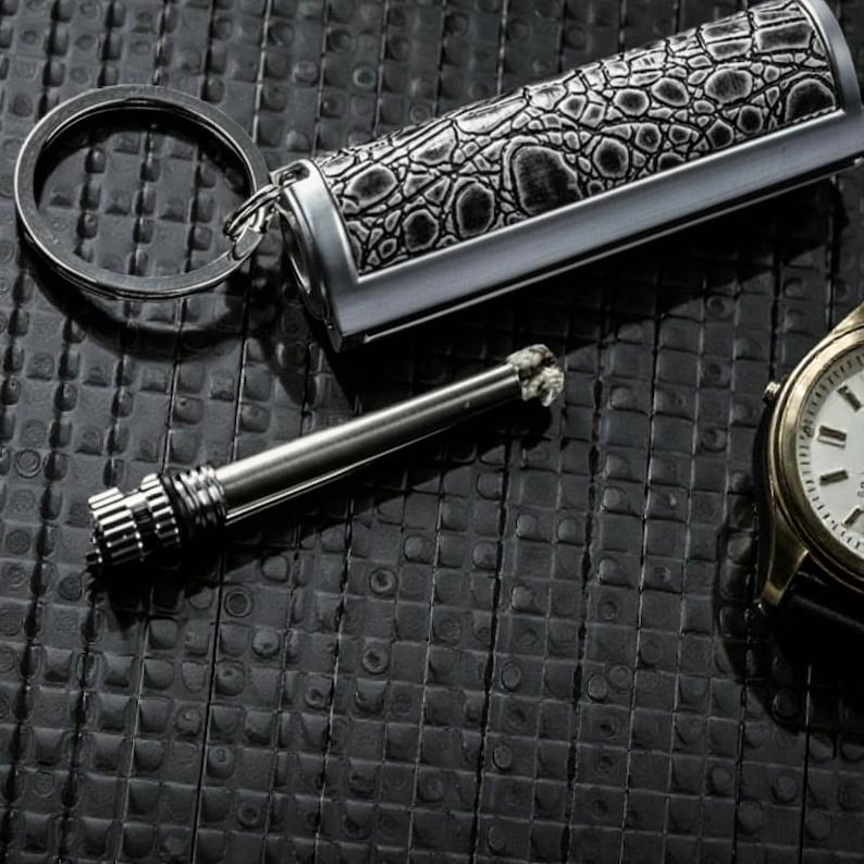 May include: A silver and black keychain lighter with a crocodile skin pattern. A separate metal striker and a gold wristwatch with a black strap are also visible. The items are on a textured black surface.