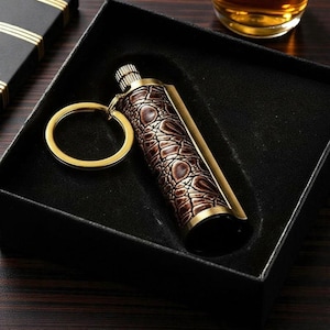 May include: A brown and gold keychain lighter with a textured, crocodile-like pattern. The lighter is in a black box, with a gold key ring attached. The lighter has a gold-coloured metal trim.