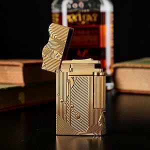 Puede incluir: Un encendedor dorado con la tapa abierta, que muestra un dise&ntilde;o grabado detallado de l&iacute;neas verticales y formas de gotas de agua. El encendedor est&aacute; sobre un fondo borroso con una botella y libros.