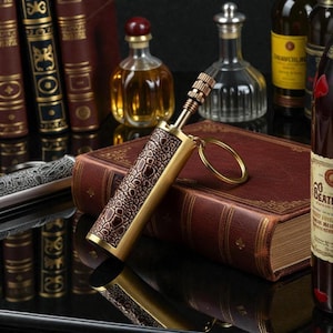 May include: A decorative brass lighter with a brown faux-leather crocodile skin pattern. The lighter has a gold keyring attached. The background includes antique books, bottles of liquor, and a knife.