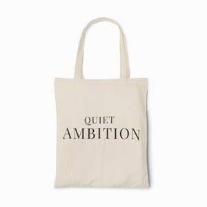 May include: A cream-colored tote bag with the words "QUIET AMBITION" printed in black. The bag has two handles and is made of a sturdy fabric, suitable for carrying various items. The design is simple and elegant.