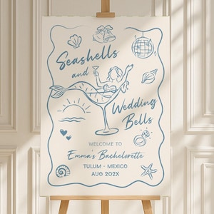 Puede incluir: Un letrero de bienvenida de color beige claro sobre un caballete de madera. El letrero presenta un dibujo lineal azul con las palabras "Seashells and Wedding Bells" y una sirena en una copa de cóctel. El letrero da la bienvenida a los invitados a la despedida de soltera de Emma en Tulum, México, agosto de 202X.