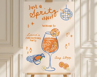 May include: A wooden easel displays a cream-colored sign with orange and blue illustrations. The sign reads "Just a Spritz older!" and features a cocktail glass, disco ball, and the text "welcome to Lucia's 25th birthday party Aug 2027."