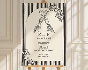 May include: A wooden easel displays a sign with a black and white striped border. The sign has the text "R.I.P. SINGLE LIFE" with skeleton hands toasting champagne flutes. Floral accents and event details are included.