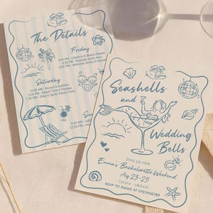 May include: Two light blue invitation cards with illustrations and text for a bachelorette weekend. One card says "The Details" and the other says "Seashells and Wedding Bells". The cards are on a white surface.