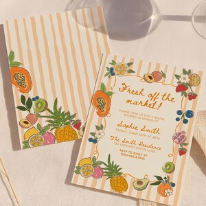 May include: Two rectangular paper cards with a light orange and white vertical stripe pattern. The cards feature colorful illustrations of various fruits including papaya, pineapple, and berries. The text reads: Fresh off the market! Please join us for a bridal shower in honor of Sophie Smith, Friday, June 15th at 6pm, The Smith Residence, 123 Orchard Grove Lane, RSVP to Emily at 123.456.87945.