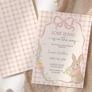 Op de afbeelding: Een uitnodiging voor een babyshower met een roze ruitpatroon en de tekst "Some Bunny is on the Way". De uitnodiging toont een cartoonkonijn met bloemen en een decoratieve bloemenrand. De uitnodiging is ongeveer 13 x 18 cm groot.