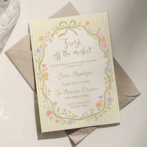 Puede incluir: Una invitación a una despedida de soltera con el texto "Fresh off the market" en una elegante escritura, rodeada por un borde floral. La invitación está sobre un fondo crema con rayas verticales e incluye detalles como la fecha, hora y lugar.