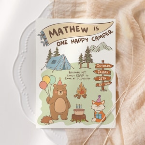 May include: A whimsical invitation card with the text "MATHEW IS ONE HAPPY CAMPER." The illustration features a bear holding balloons, a fox with a gift, a tent, a campfire, and a birthday cake. The card includes RSVP details and event information.