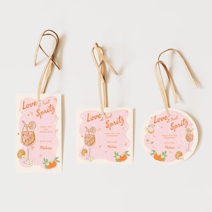 May include: Three gift tags with a pink background and the text "Love at First Spritz". Each tag features a cocktail illustration, orange accents, and a raffia ribbon. The tags are in rectangular, square, and circular shapes.