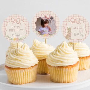 May include: Cupcakes with white frosting on a white plate. The cupcakes have decorative toppers with text that reads "Happy Birthday Alisa!" and "Alisa's 1st Birthday". One topper has a photo of a child.