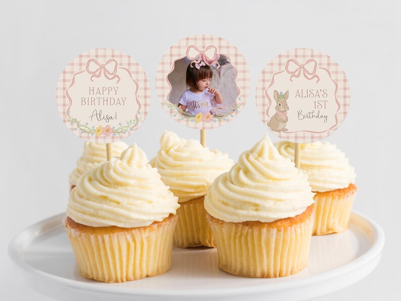 May include: Cupcakes with white frosting on a white plate. The cupcakes have decorative toppers with text that reads "Happy Birthday Alisa!" and "Alisa's 1st Birthday". One topper has a photo of a child.