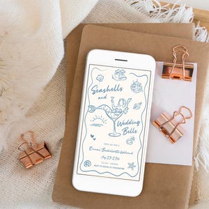 May include: A smartphone screen shows a light blue invitation with the words "Seashells and Wedding Bells." The design includes a mermaid in a cocktail glass and other illustrations. Copper binder clips and stationery are also visible.