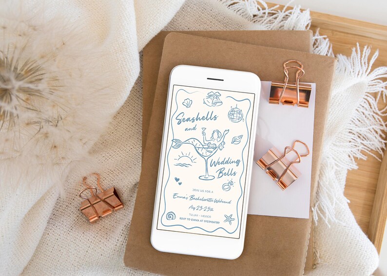 May include: A smartphone screen shows a light blue invitation with the words "Seashells and Wedding Bells." The design includes a mermaid in a cocktail glass and other illustrations. Copper binder clips and stationery are also visible.