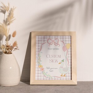 May include: A framed custom sign with a light wood frame. The sign features a cartoon goose with balloons and the text "Custom Sign". A vase with dried flowers is to the left.
