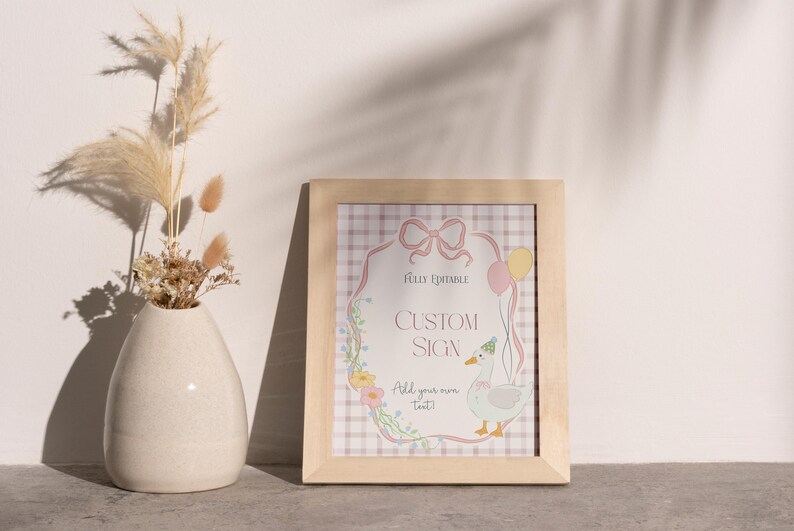 May include: A framed custom sign with a light wood frame. The sign features a cartoon goose with balloons and the text "Custom Sign". A vase with dried flowers is to the left.