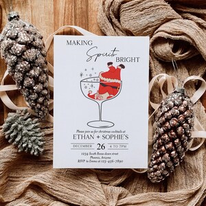 May include: A Christmas cocktail invitation with the text "MAKING Spirit BRIGHT". The invitation features a Santa Claus illustration in a cocktail glass. Two pinecone ornaments and fabric complete the festive scene.