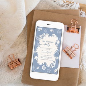 May include: A white smartphone displays a baby shower invitation with a blue and cream seashell design. The invitation reads "Sea You Soon baby" and includes event details. Two rose gold binder clips and a stack of brown paper are also visible.