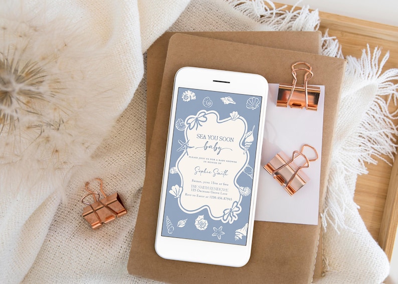 May include: A white smartphone displays a baby shower invitation with a blue and cream seashell design. The invitation reads "Sea You Soon baby" and includes event details. Two rose gold binder clips and a stack of brown paper are also visible.