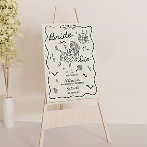Pode incluir: Um cavalete de madeira clara exibe uma placa com as palavras "Bride or Die" em uma fonte extravagante. A placa apresenta uma ilustra&ccedil;&atilde;o em preto e branco de uma mulher em um copo de martini, com texto adicional e elementos decorativos. A placa &eacute; para um fim de semana de despedida de solteira.