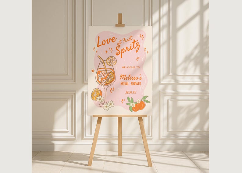 May include: A wooden easel displays a sign with the words "Love at First Spritz" in orange script. The sign features a cocktail illustration with orange slices and the text "Melissa's Bridal Shower". The background is a light pink shape with orange accents.
