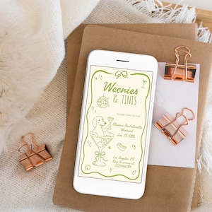 May include: A smartphone displays a light green invitation with the text "Weenies & Tinis" and a cartoon dog in a martini glass. The phone rests on brown paper with rose gold binder clips. A white cloth and wooden tray are in the background.