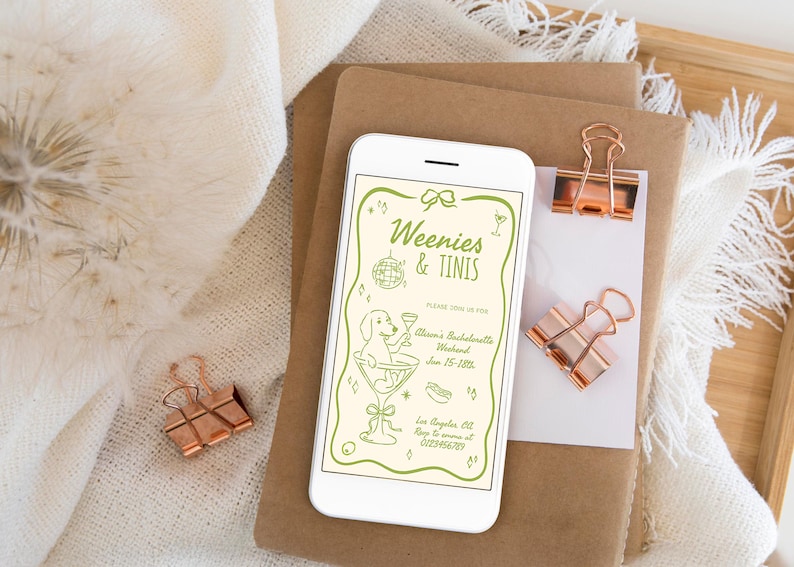 May include: A smartphone displays a light green invitation with the text "Weenies & Tinis" and a cartoon dog in a martini glass. The phone rests on brown paper with rose gold binder clips. A white cloth and wooden tray are in the background.
