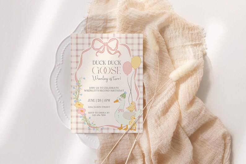 May include: A pastel-colored birthday invitation with the text "Duck Duck Goose Wrenley is two!" features a cartoon goose and ducklings. The invitation is on a white plate with a peach-colored fabric in the background.