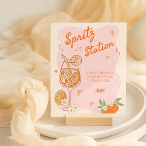 以下が含まれることがあります： オレンジ色のスクリプトで「Spritz Station」という言葉が書かれた正方形のサイン。オレンジのスライスが入ったカクテルのイラストが特徴です。サインにはレシピが含まれており、木製のスタンドに表示されています。
