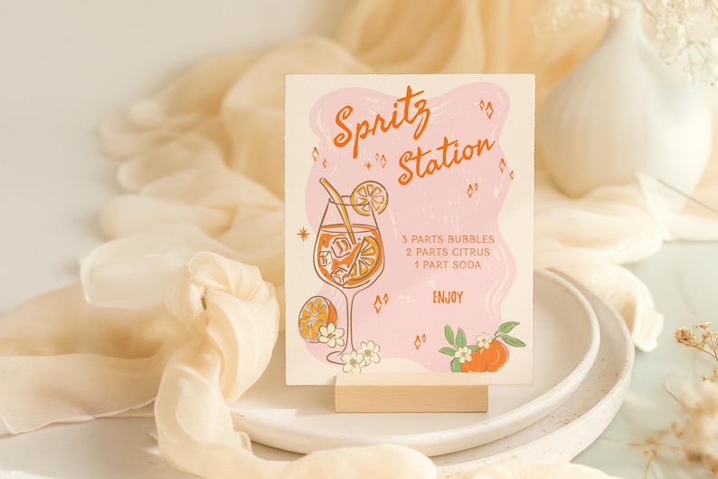 以下が含まれることがあります： オレンジ色のスクリプトで「Spritz Station」という言葉が書かれた正方形のサイン。オレンジのスライスが入ったカクテルのイラストが特徴です。サインにはレシピが含まれており、木製のスタンドに表示されています。
