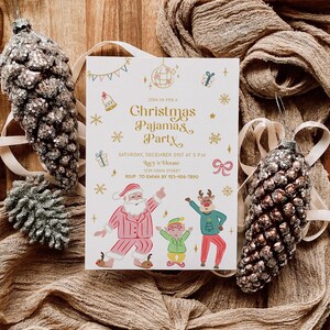 Op de afbeelding: Een uitnodigingskaart voor een kerstpyjamaparty met de kerstman, een rendier en een elf die dansen. De uitnodiging heeft gouden tekst en feestelijke illustraties op een witte achtergrond. Twee glinsterende dennenappelornamenten en beige stof omringen de kaart.