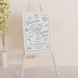 May include: A wooden easel supports a sign with blue illustrations and text. The sign says "Seashells and Wedding Bells" with a mermaid in a cocktail glass. Additional text welcomes guests to Emma's Bachelorette in Tulum, Mexico.
