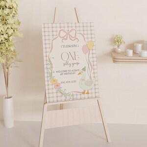 May include: A pink and white sign with a gingham pattern, featuring a cartoon goose with a party hat and the text "Celebrating One Silly Goose. Welcome to Alma's 1st Birthday. June 16th, 2024."