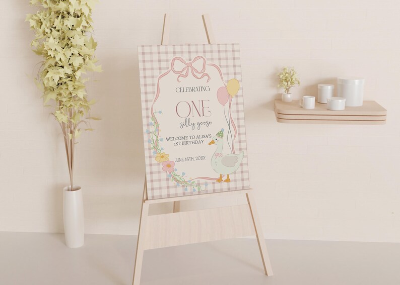 May include: A pink and white sign with a gingham pattern, featuring a cartoon goose with a party hat and the text "Celebrating One Silly Goose. Welcome to Alma's 1st Birthday. June 16th, 2024."