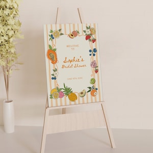 May include: A rectangular sign on a wooden easel featuring a cream background with vertical light orange stripes. The border is decorated with colorful illustrations of various fruits. The text reads: WELCOME TO Sophie's Bridal Shower JUNE 15TH, 20XX.