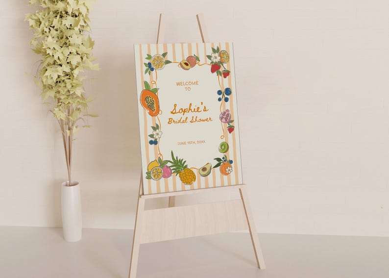 May include: A rectangular sign on a wooden easel featuring a cream background with vertical light orange stripes. The border is decorated with colorful illustrations of various fruits. The text reads: WELCOME TO Sophie's Bridal Shower JUNE 15TH, 20XX.