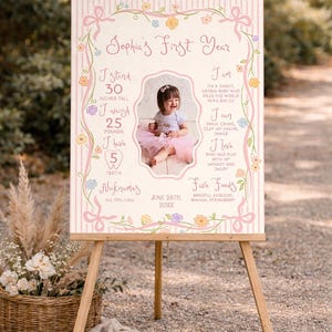 May include: A first birthday sign on an easel, decorated with pink ribbons, flowers, and a photo of a baby girl. The sign states the baby is 76 cm tall, weighs 11 kg, and has 5 teeth. The sign includes the baby's nicknames.