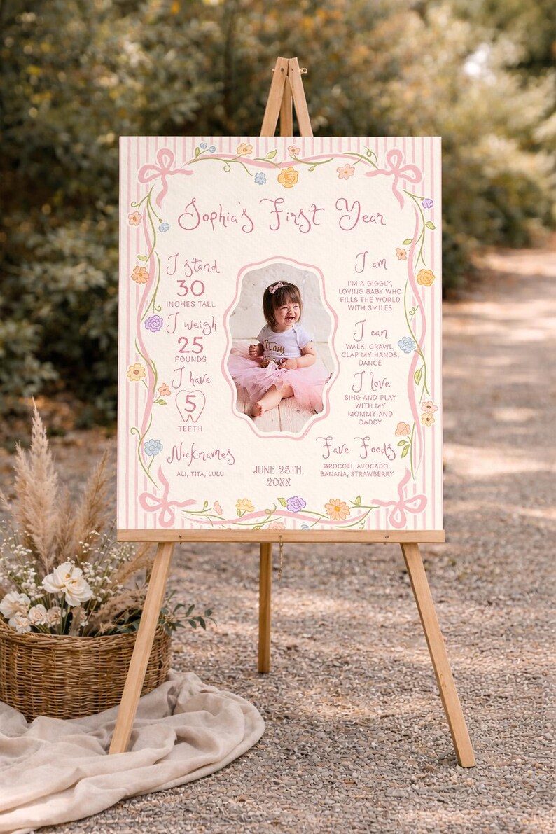 May include: A first birthday sign on an easel, decorated with pink ribbons, flowers, and a photo of a baby girl. The sign states the baby is 76 cm tall, weighs 11 kg, and has 5 teeth. The sign includes the baby's nicknames.
