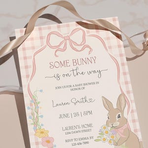 Op de afbeelding: Een uitnodiging voor een babyshower met de tekst "SOME BUNNY is on the way". De uitnodiging heeft een roze ruitpatroon, een roze lint en een cartoonkonijn met bloemen. Extra tekst bevat de naam van de ge&euml;erde, datum, tijd en adres.
