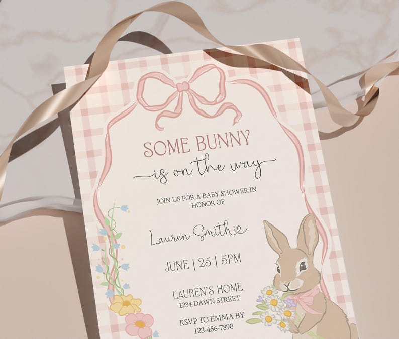 Op de afbeelding: Een uitnodiging voor een babyshower met de tekst "SOME BUNNY is on the way". De uitnodiging heeft een roze ruitpatroon, een roze lint en een cartoonkonijn met bloemen. Extra tekst bevat de naam van de ge&euml;erde, datum, tijd en adres.