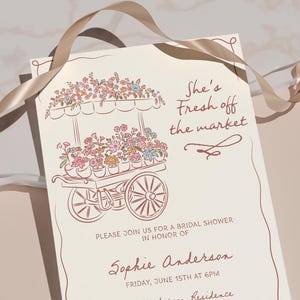 May include: Cream-colored invitation with a floral cart illustration in brown ink. Text reads "She's Fresh off the market" and details for a bridal shower. A tan ribbon is draped across the top.