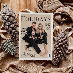 May include: A magazine cover with the text "HAPPY HOLIDAYS 2025 FAMILY EDITION" featuring a family portrait. The magazine is surrounded by decorative pinecone ornaments and draped fabric, creating a festive holiday theme.